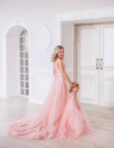 妈妈和女儿穿着豪华的粉红色连衣裙和一列长长的火车。 家庭服装相同的衣服。 背景是美丽昂贵的白色经典内饰。 艺术家庭照片