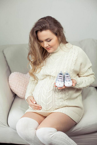 一个年轻漂亮的孕妇抚摸着她的肚子。一个穿着针织连衣裙的孕妇坐在沙发上, 抱着孩子的鞋子。母亲.怀孕.一个快乐的孕妇的肖像。舒适
