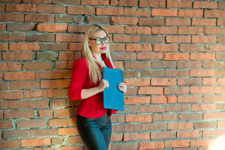 一个年轻漂亮的女孩穿着红色夹克，戴着眼镜，站在砖墙旁边，手里拿着一个文件夹