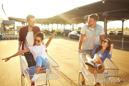 一群快乐的年轻人在购物车上玩得很开心。 多民族年轻人在购物车上赛车。 阳光明媚的夏日。 生活方式的概念。 一群朋友享受生活。