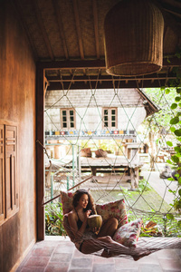 女人坐在吊床上，抱着五颜六色的枕头，喝着新鲜的年轻椰子。 乡村风格的木屋