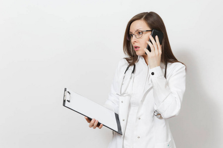 严重的年轻医生与听诊器, 眼镜被隔离在白色背景。女医生在医疗礼服上讲手机, 在记事本文件夹上保存健康卡。医护人员理念