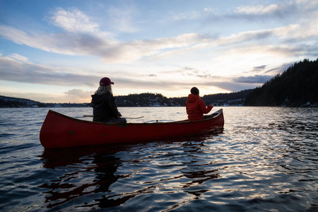 在一个充满活力的日落中，一对坐在木制独木舟上的朋友正在一个被加拿大山脉包围的入口划水。 在加拿大温哥华北深海湾附近的印度手臂上。
