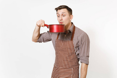 年轻的乐趣疯狂的人厨师或服务员在条纹棕色围裙, 衬衫品尝从红色空 stewpan 在头被隔绝在白色背景。男管家或 housewo
