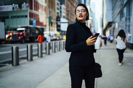 穿着时髦的女经理拿着智能手机查看邮件，漫步在曼哈顿街头，美丽的女商人带着红唇和眼镜，带着咖啡走着发送信息