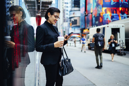自信的女主人，公司漫步与咖啡，去发送反馈的智能手机站在市中心。