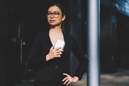 有经验的女经济学家穿着时尚的黑色西装和光学眼镜，手里拿着现代数字手机，站在办公楼的宣传背景上看着。