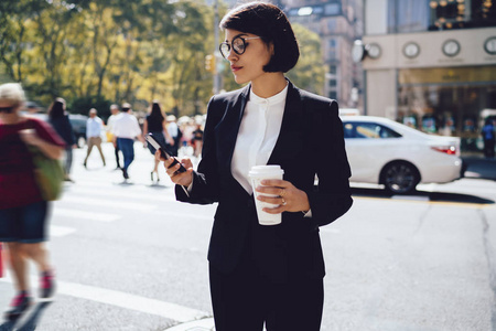 自信的女企业家用手机发信息，站在纽约拥挤的道路上，严肃的女商人用手机在网页上阅读信息，手里拿着模拟咖啡杯