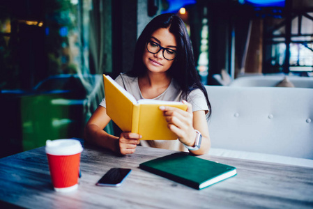 带着眼镜的沉思的年轻学生在咖啡休息时间读最喜欢的书，花时间在自编自演，专心的女商人在咖啡馆的空闲时间检查牛奶场的工作日程
