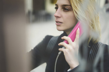 迷人的金发女性旅行者在户外使用公共电话付费连接，年轻的时髦女孩在电话亭拨打电话，站在模糊的复制空间区域附近做广告