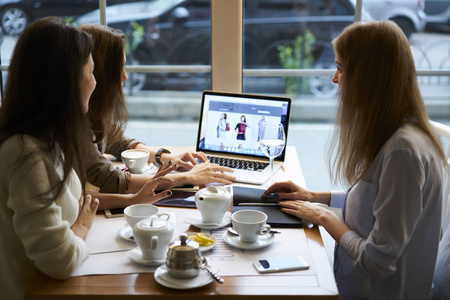 女商务同事浏览网上商店网页的后视图与新服装收藏，合作伙伴在咖啡厅茶点交流分享共同项目的意见