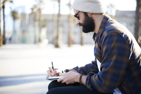 休闲装和时尚太阳镜中的英俊胡子男作者在记事本中创作了一篇关于坐在长凳上享受春天天气的文章，在你的广告复制空间区域附近