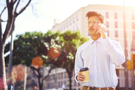 英俊的严肃的男性游客在酷眼镜使用电话应用进行移动对话。年轻人在阳光街上散步时通过智能手机与朋友交流