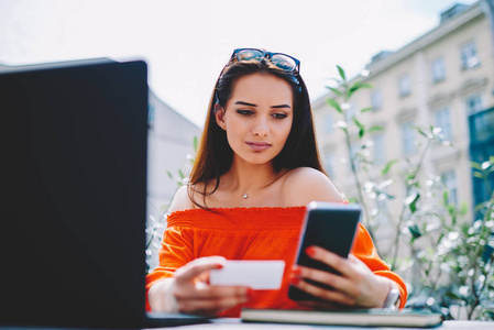 一位年轻的黑发女人，头上戴着眼镜，在互联网网站上寻找新工作，在智能手机设备上拨打电话号码，名片坐在市中心
