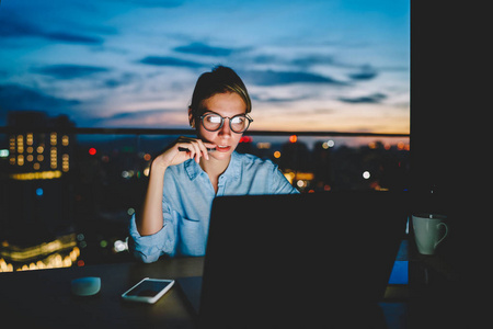 深夜家漂亮的思考女企业家在酷眼镜中通过笔记本电脑上的应用程序与