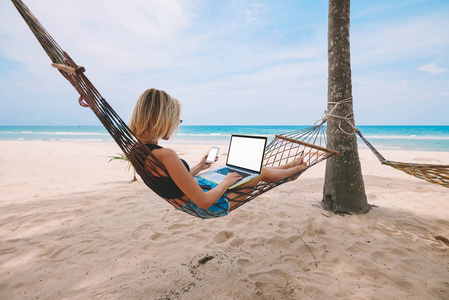 女性自由职业者企业家躺在吊床上，在现代笔记本电脑上做远程商务工作，暑假期间在热带海滨为智能手机上的网页提供空白屏幕区域