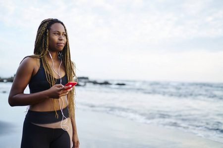 运动的黑人女性慢跑者穿着活跃的服装，听着耳机里的音乐，在早上的海岸上与电话相连