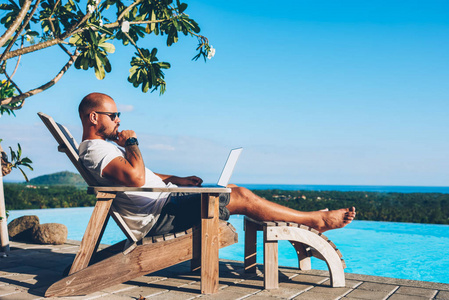 一位留着胡子的商人躺在蓝色游泳池附近，一边在上网本的键盘上打字，屏幕上有空白的复制空间，在热带地区的避暑胜地进行远程广告工作