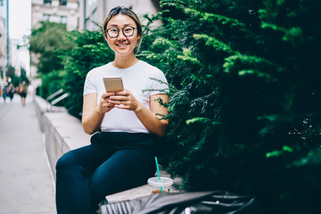积极向上的亚洲年轻女子在市中心休息，与朋友在线聊天，在移动，快乐的中国女孩戴眼镜，在社交网络中分享多媒体文件，在市中心休闲。