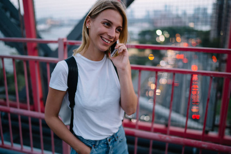 兴高采烈的时髦女孩，喜欢用良好的移动连接进行电话交谈，走在桥上，微笑的女游客在漫游中用智能手机交谈，通讯费用低廉