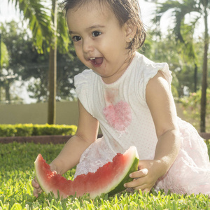 两岁的女孩穿着白色漂亮的粉红色连衣裙，坐在绿色的草头上，头上戴着他手里拿着的西瓜或红瓜。 玩笑笑搞笑搞笑表情