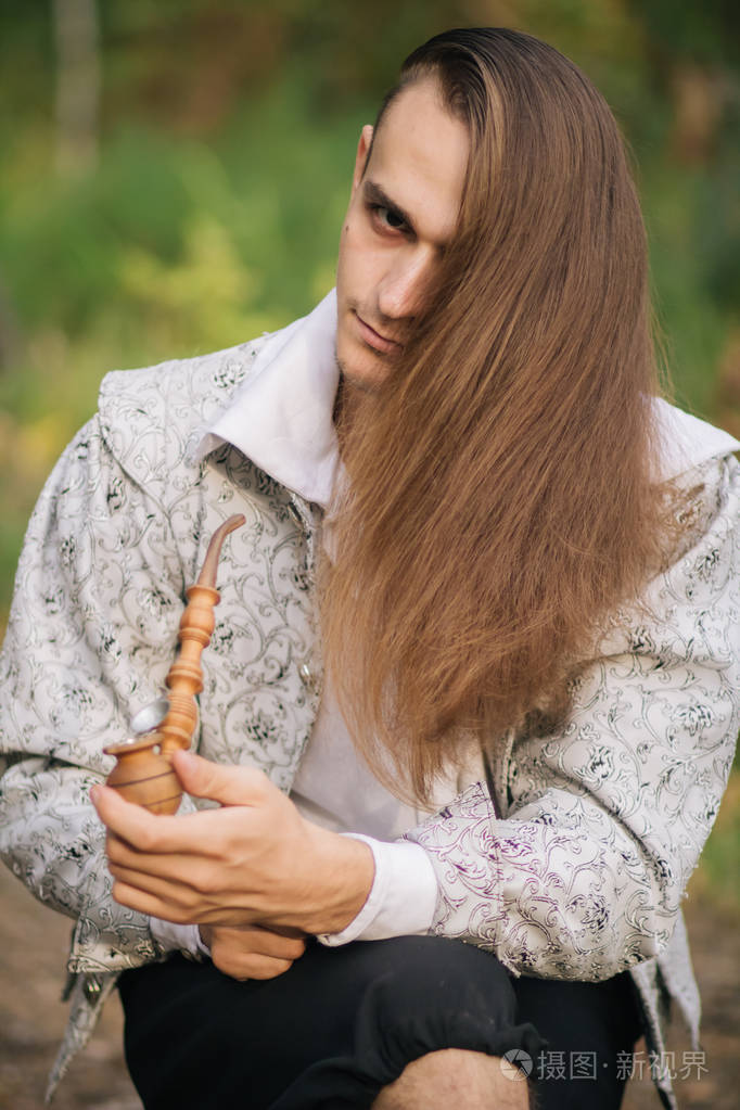 性感的长发男人,手里拿着烟斗在树林里.