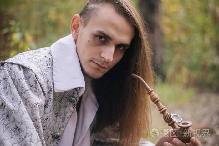 性感的长发男人手里拿着烟斗在树林里穿着一件漂亮的古代夹克有图案和