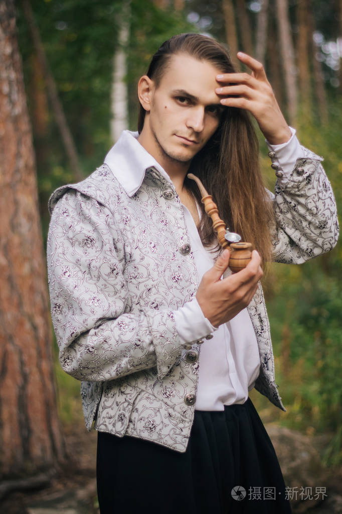 性感的长发男人手里拿着烟斗在树林里穿着一件漂亮的古代夹克有图案和