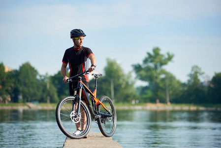 运动员骑自行车的职业自行车服装和防护头盔摆在自行车附近，看着很远的思考未来的成功和成就。 运动员在湖边的码头休息。