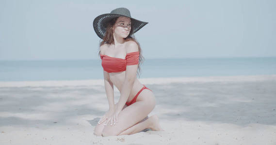 美丽的女孩，穿着比基尼和帽子，在暑假在阳光下放松，在热带沙滩上的海洋和天空背景。