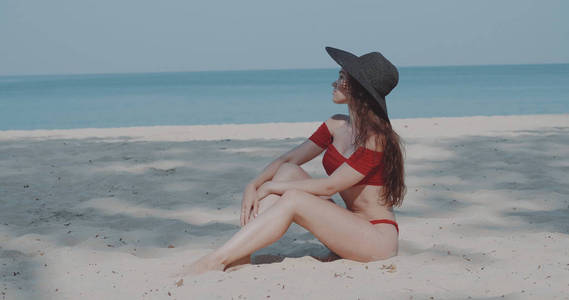 美丽的女孩，穿着比基尼和帽子，在暑假在阳光下放松，在热带沙滩上的海洋和天空背景。
