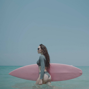 年轻快乐的女人，穿着泳衣和太阳镜，在阳光明媚的夏日带着粉红色的冲浪板走向大海。 美丽的女孩享受热带海滩假期