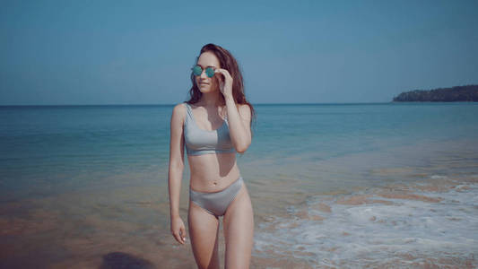 美丽快乐的年轻女孩穿着比基尼和太阳镜从海水中走出来。 美丽的女人站在天空背景下的热带海滩上