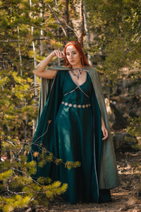 秋天的森林里走着一个美丽的红发女孩，她穿着一件绿松石的连衣裙，头上戴着一顶。在精灵的形象里。它在山上的大岩石上。太阳眩光。幻想