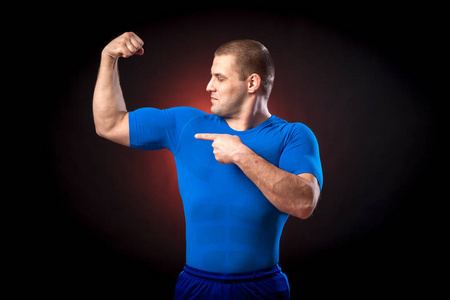 一个穿着蓝色运动服的强壮的深色头发的运动员，带着匆忙的微笑，在黑色的孤立背景上展示二头肌与红色的灯光