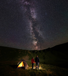 后景年轻夫妇游客牵手欣赏星空和迷人明亮的银河。 在湖边的山上露营时，男人和女人在篝火旁休息。