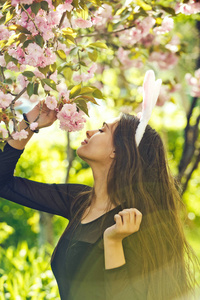 女孩或漂亮的女人，玫瑰色的兔子耳朵和长长的黑发闻着樱花花从春天公园在阳光明媚的日子在模糊的花卉环境。 复活节。 春季