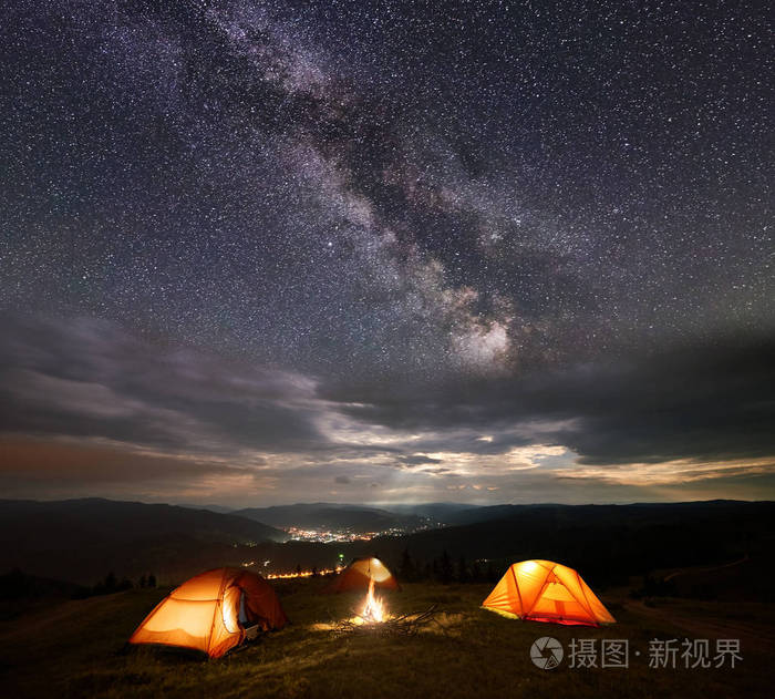在星空下的夜晚,三顶照亮的橙色帐篷在山上,银河和云在山谷中发光的
