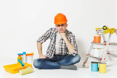 严肃的人在橙色防护头盔坐在地板上的装修设备的房间, 在白色背景隔离。墙纸胶水配件, 喷漆工具。修复主页概念