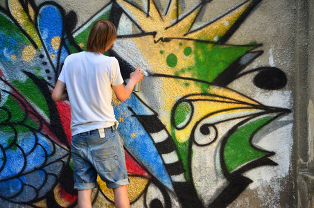 一个穿牛仔短裤和白衬衫的年轻人的照片。 这家伙在涂鸦墙上画了一幅用各种颜色的气溶胶画的画。 流氓和破坏财产的概念