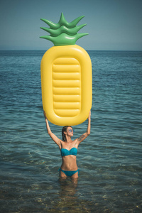 妇女在自己之上举行空气床垫菠萝形状, 海或海洋在背景。暑假的概念。女士与空气床垫站在水中, 穿着时髦的比基尼和太阳镜