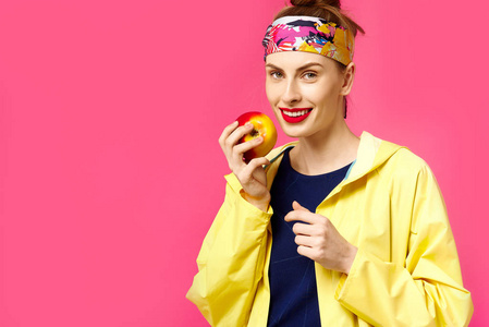 一个粉红色的背景和黄色的大衣的年轻女子在她手中持有一个苹果。健康营养和体育的概念。色彩痴迷的概念。简约风格。时尚时尚