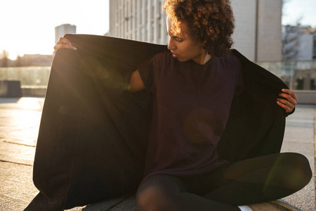 冷卷的非裔美国女孩坐在洛托斯的姿势在屋顶上，穿上羊毛衫，日落背光的城市景观。 清晨在日出的城市背景下卷曲的女人