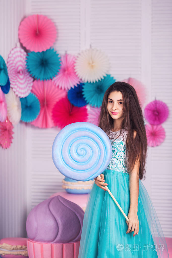 美丽的女孩在蓝色礼服手拿着巨大的糖果在工作室与装饰的杏仁复制空间