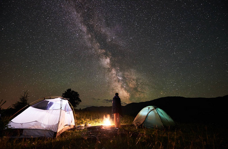 活跃的女徒步旅行者晚上在山上休息，在充满星星和银河的惊人夜空下露营。女游客站在篝火旁和两顶照明帐篷后景