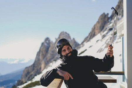 胡子微笑 snowboarded 在太阳镜面具做自拍在智能手机在滑雪胜地。模糊的山和蓝天的背景。水平图像