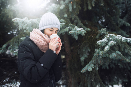 美丽的年轻白种女人在冬季夹克, 针织豆帽灰色帽子和粉红色的围巾, 拿着一杯咖啡或茶, 在户外公园或森林在下雪的冬季天。冬季心情