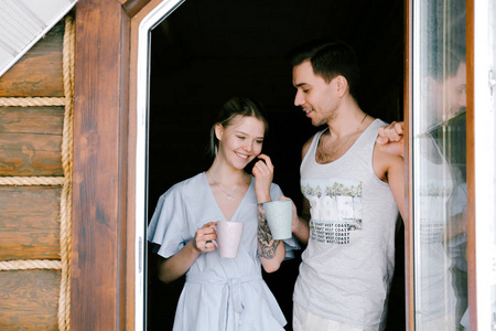 可爱的夫妇在家在一起。 漂亮的女人和迷人的男人穿着时尚的休闲服站在阳台上喝茶笑。