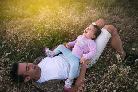 英俊的年轻父亲, 他可爱的蹒跚学步的女孩在草地上玩耍和笑。快乐的父亲与他的女婴在一个晴朗的一天或晚上。家庭漫步在草地上