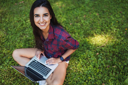 年轻白种黑发妇女的顶部视图微笑着穿着格子衫, 坐在笔记本电脑前的草地上, 用无线通讯购物和学习的笔记本电脑工作。